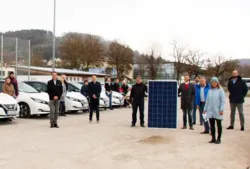 Menschen stehen auf einem Parkplatz, wo Autos zu sehen sind. Außerdem halten zwei Personen eine Solarzelle.