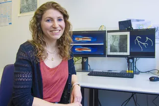 Eine Frau sitzt vor ihrem Computer und schaut in die Kamera. Im Hintergrund sind zwei Bildschirme mit Simulationen von Herzkranzgefäßen